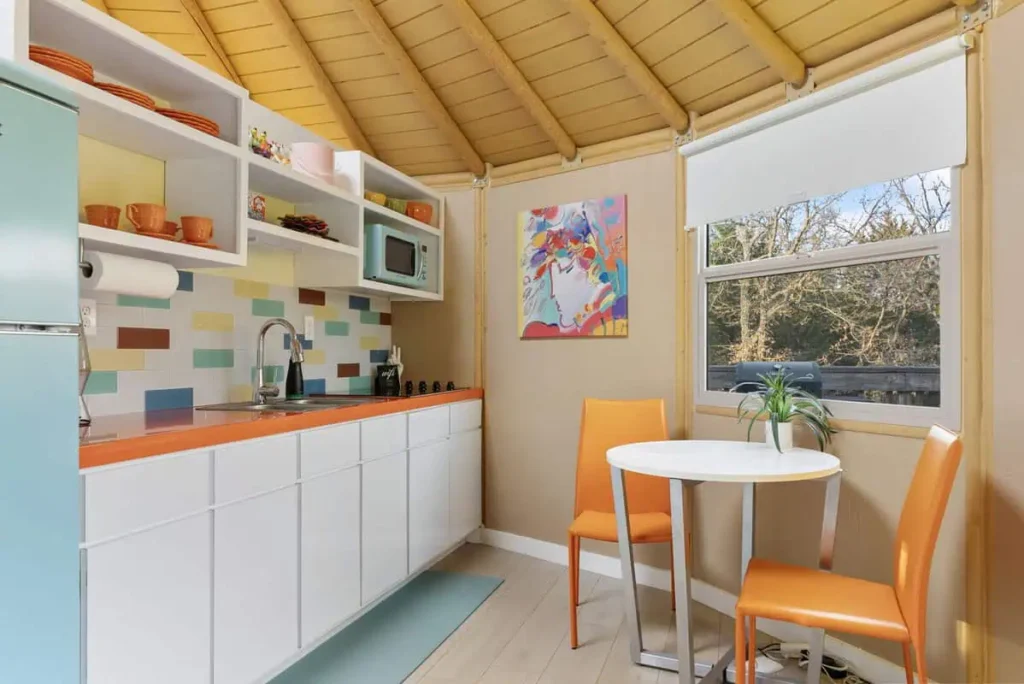 Cozy kitchen interior of the 'Hippy Chick' yurt at All Is Well Resort, featuring colorful decor, a refrigerator, microwave, and dining table with orange chairs, surrounded by natural light and whimsical art.