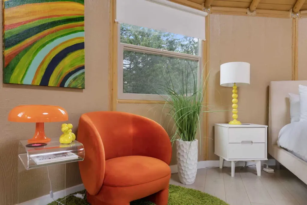 Cozy interior of the 'Hippy Chick' yurt featuring an orange chair, colorful abstract wall art, a modern lamp, and a white nightstand, highlighting the unique glamping experience at All Is Well Resort.